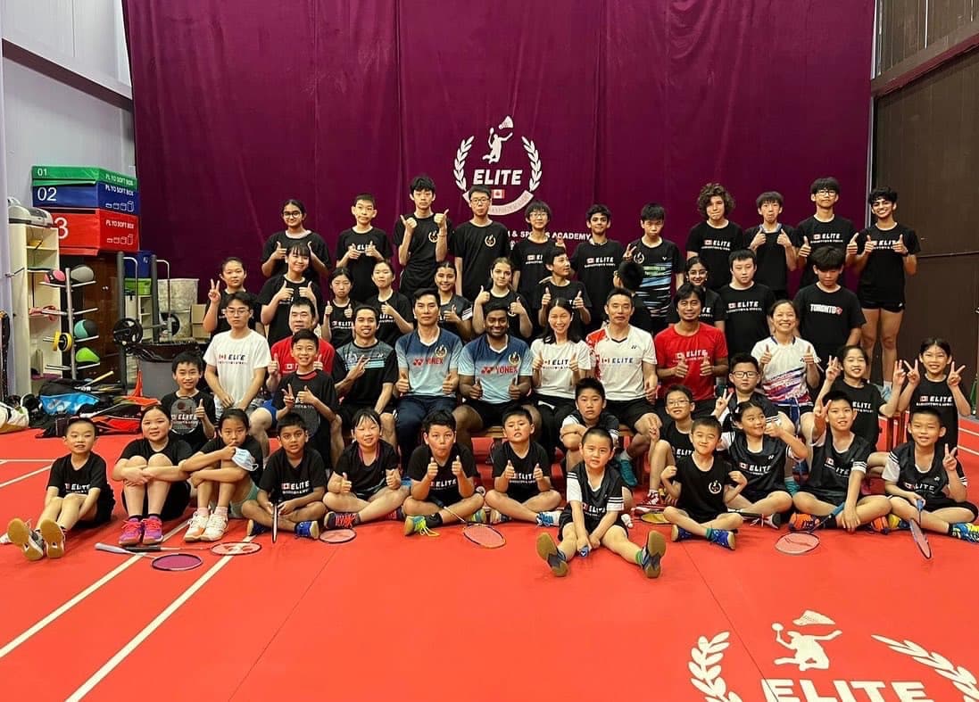 A large group of Elite Badminton coaches and students in club shirts, posing together on court in front of the Elite Badminton Academy backdrop.